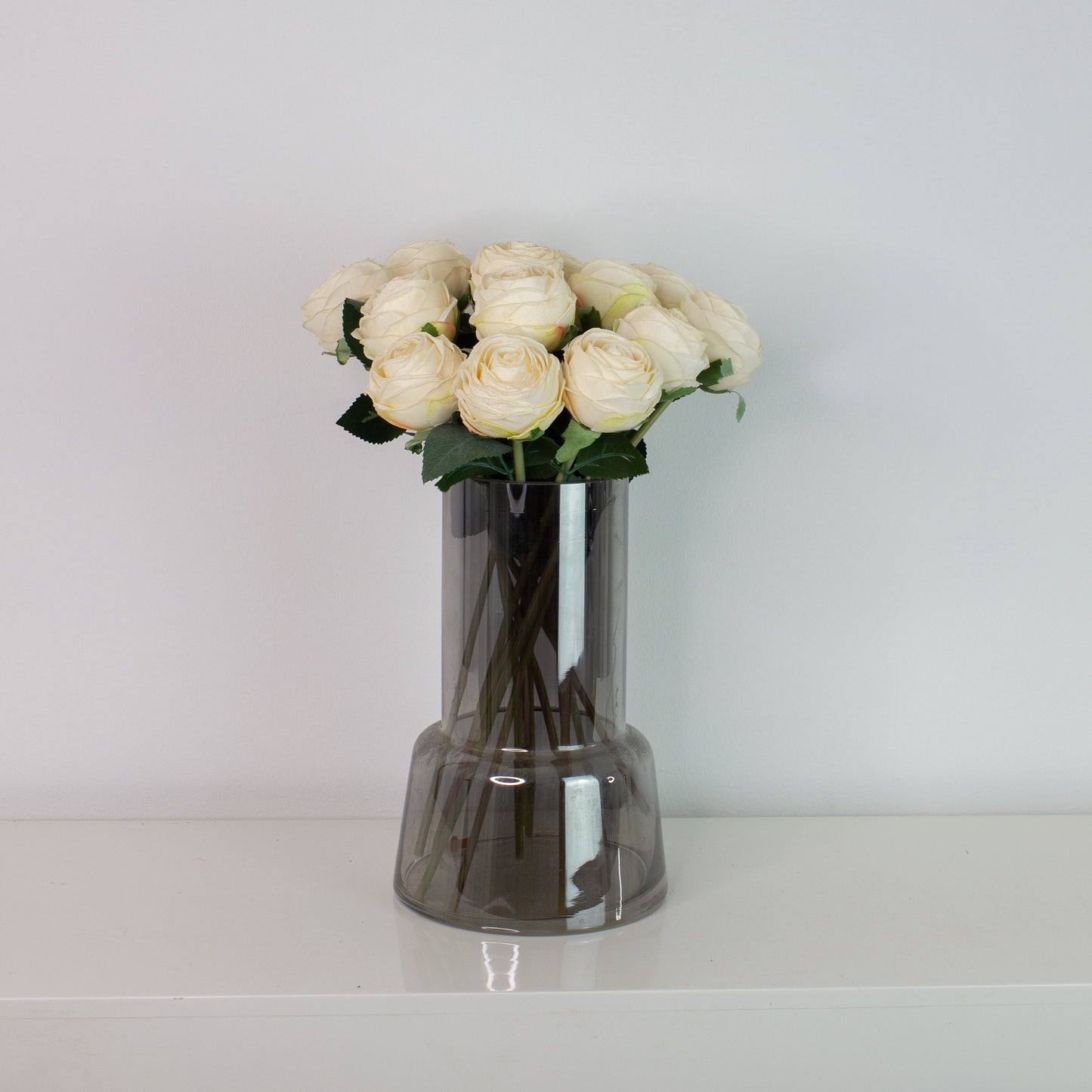 Naturtro kunstig buket med små hvide roser i mørk glasvase på hvid overflade – elegant og vedligeholdelsesfri blomsterdekoration.