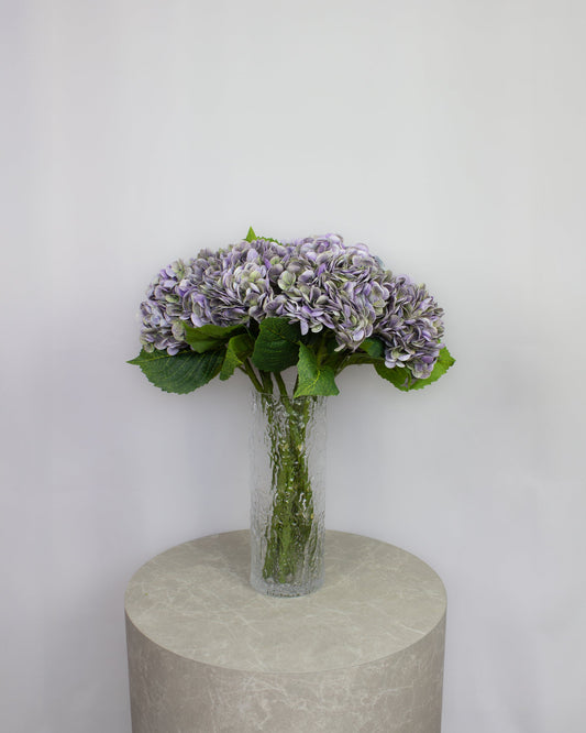 mørkelilla hortensia – naturtro kunstig buket med 9 fyldige hortensiahoveder i glasvase på bord.