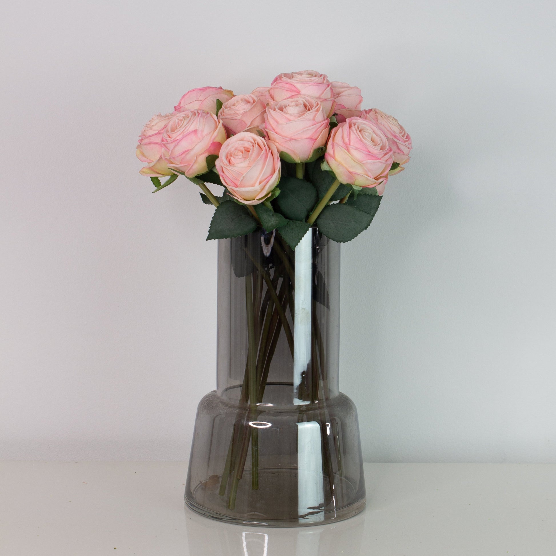 Naturtro kunstig buket med små lyserøde roser i mørk glasvase på hvid overflade – elegant og vedligeholdelsesfri blomsterdekoration.
